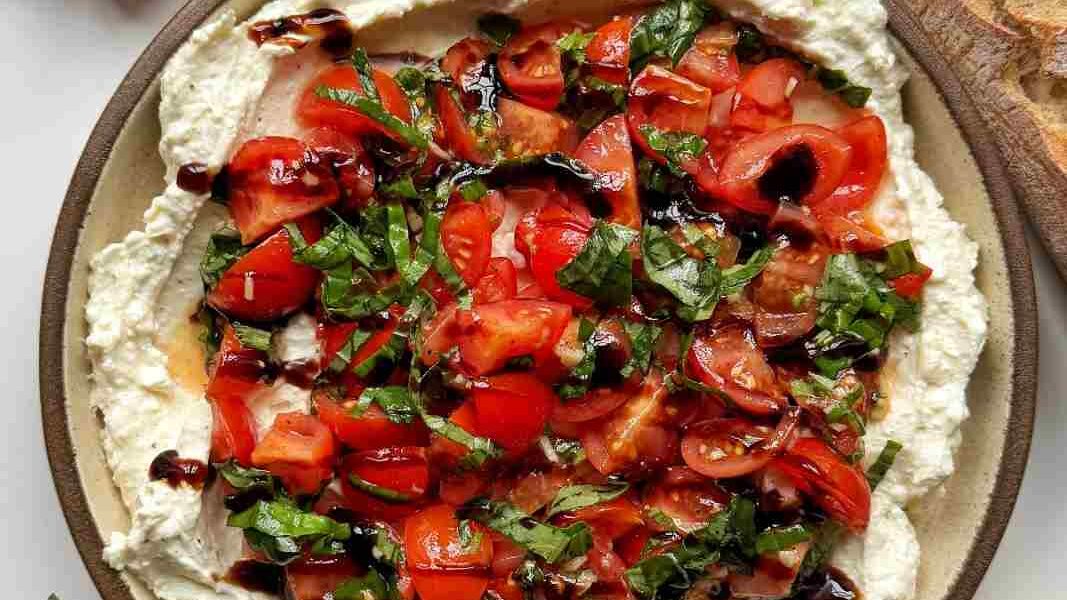 A bowl of bruschetta topping made from creamy whipped cream cheese and feta topped with garlicky basil tomatoes with olive oil and a balsamic glaze. Served up with toasted slices of baguette.