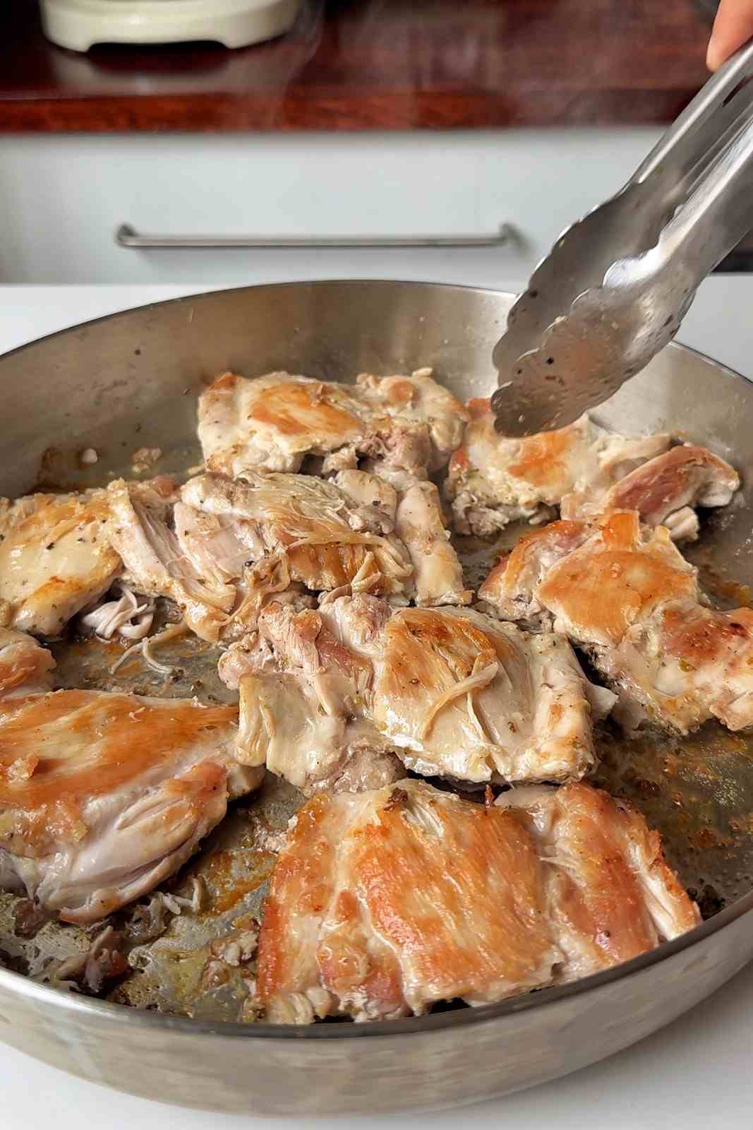 Golden seared chicken thighs in a stainless steel skillet being turned with tongs.