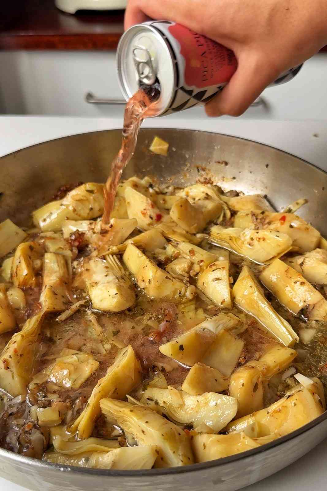 Pouring white wine from a can into the skillet of artichoke hearts.