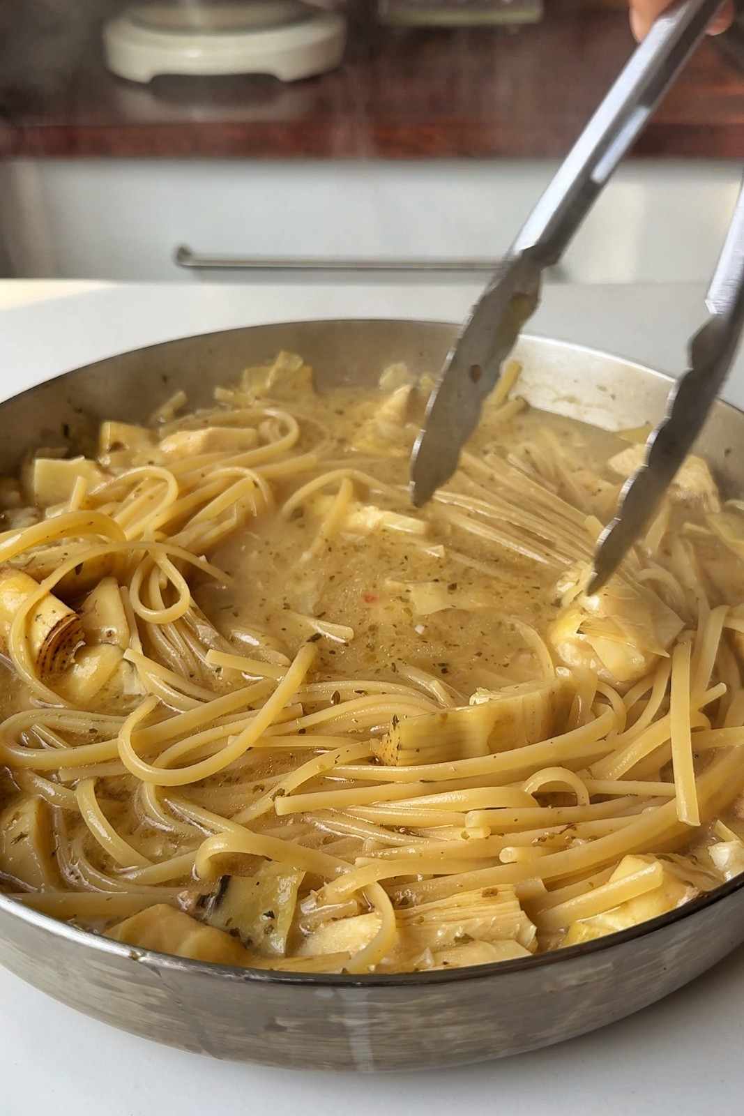 Tongs stirring linguine and artichoke hearts as they simmer in cloudy chicken stock.