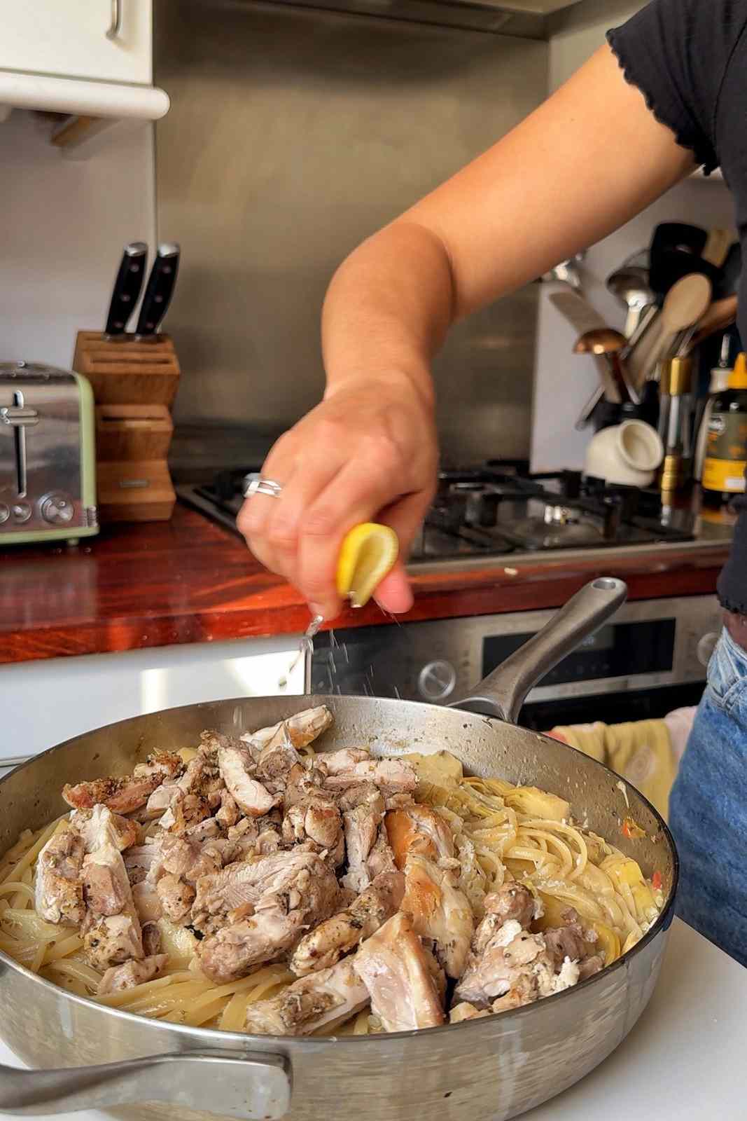 Emily Roz squeezing a lemon wedge over sliced seared chicken piled on linguine and artichokes in the skillet.