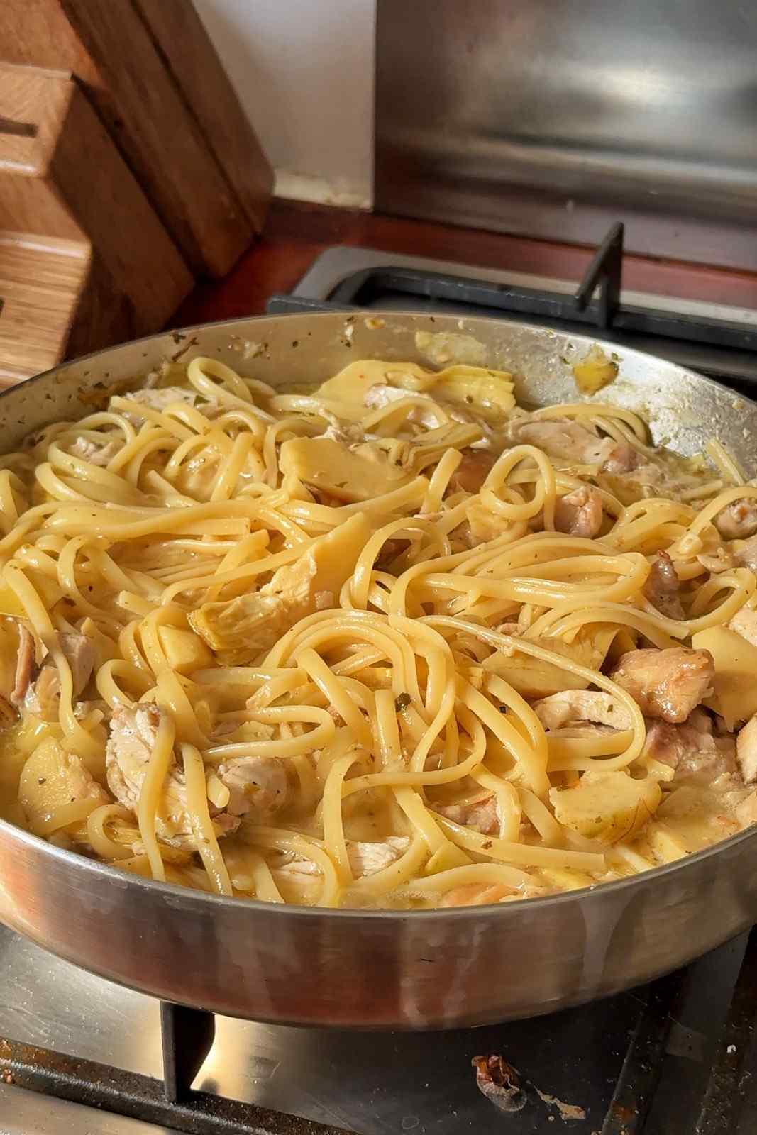 Finished chicken carciofi pasta in a skillet on the stove, with linguine, chicken and artichoke hearts in a glossy sauce.