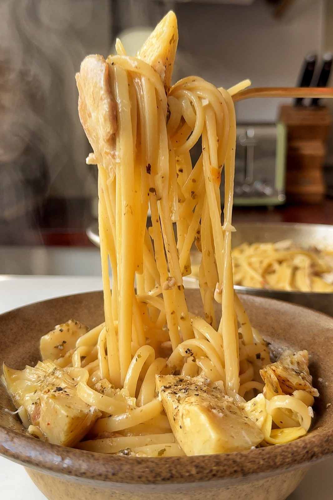 A twirl of chicken carciofi pasta lifted on a fork above a stoneware bowl, with steam rising.