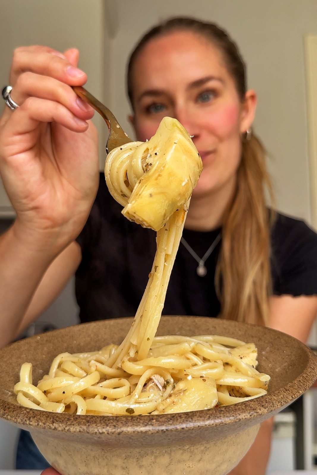 Emily holding up a forkful of linguine and an artichoke heart over a bowl of chicken carciofi pasta.