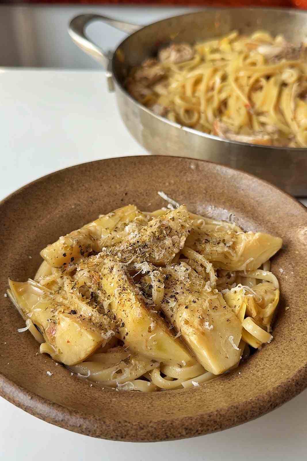 A bowl of chicken carciofi pasta topped with artichoke hearts, grated parmesan, dried oregano and black pepper, with the skillet behind.