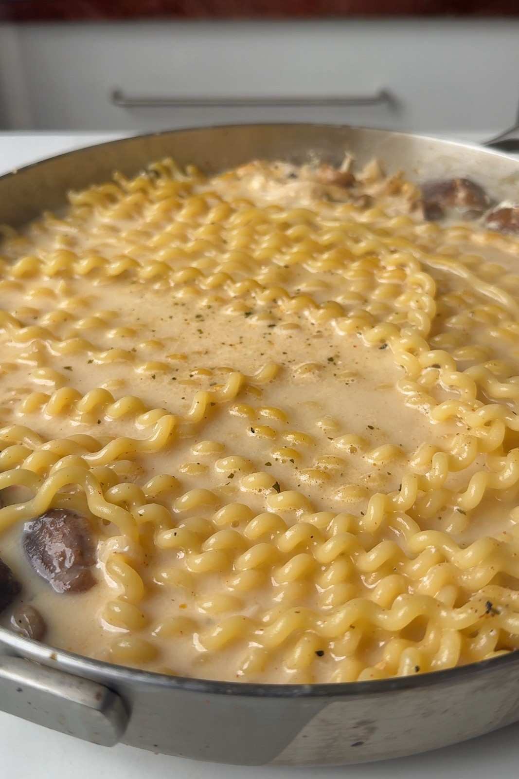 Long spiral pasta simmering in creamy vegetable stock with mushrooms in a stainless steel skillet.