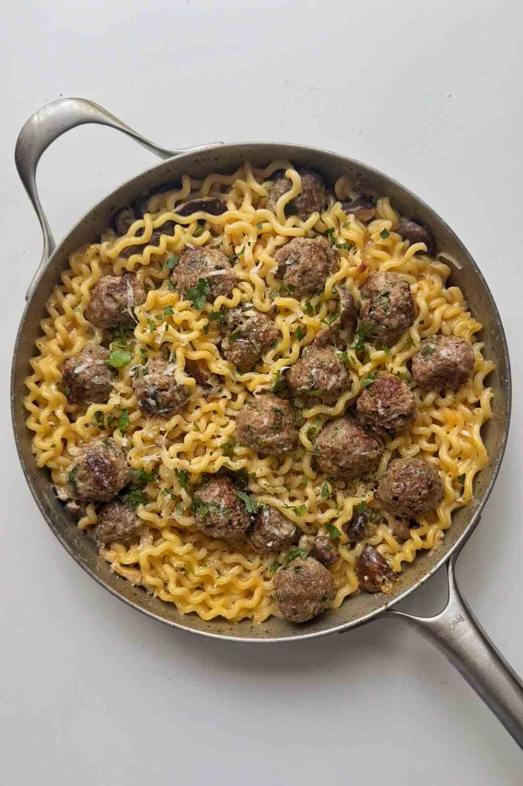 Overhead view of a skillet of creamy mushroom meatball pasta finished with grated parmesan and chopped parsley.