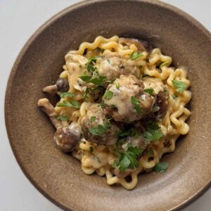 Overhead view of a bowl of long spiral pasta and pork meatballs in creamy mushroom sauce, topped with parsley.