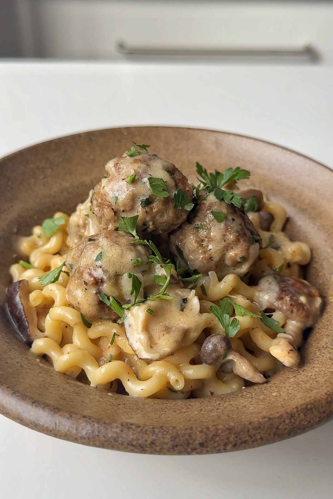 Creamy mushroom meatball pasta in a stoneware bowl, with pork meatballs, torn mushrooms and fresh parsley on top.