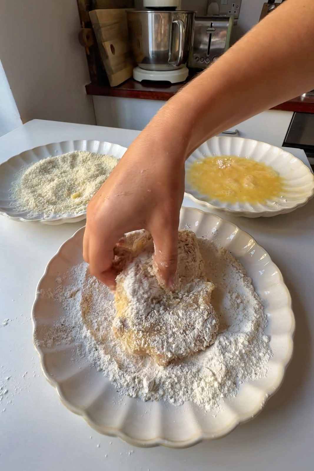 Hand coating a chicken thigh in seasoned flour, with plates of breadcrumbs and whisked egg set up behind.