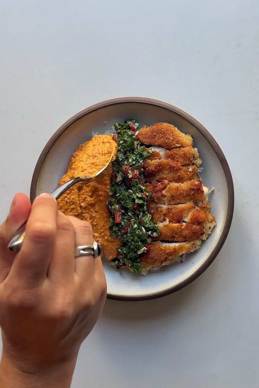 Spooning romesco sauce onto a plate of sliced crispy chicken, jasmine rice and green sauce.