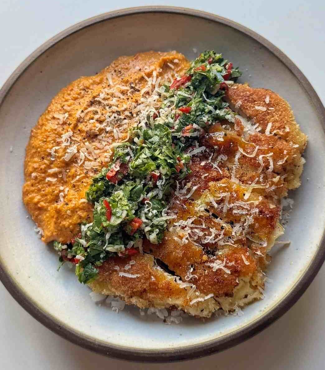 Crispy romesco chicken over jasmine rice in a stoneware bowl, with romesco sauce, a cilantro and chili green sauce and grated parmesan.