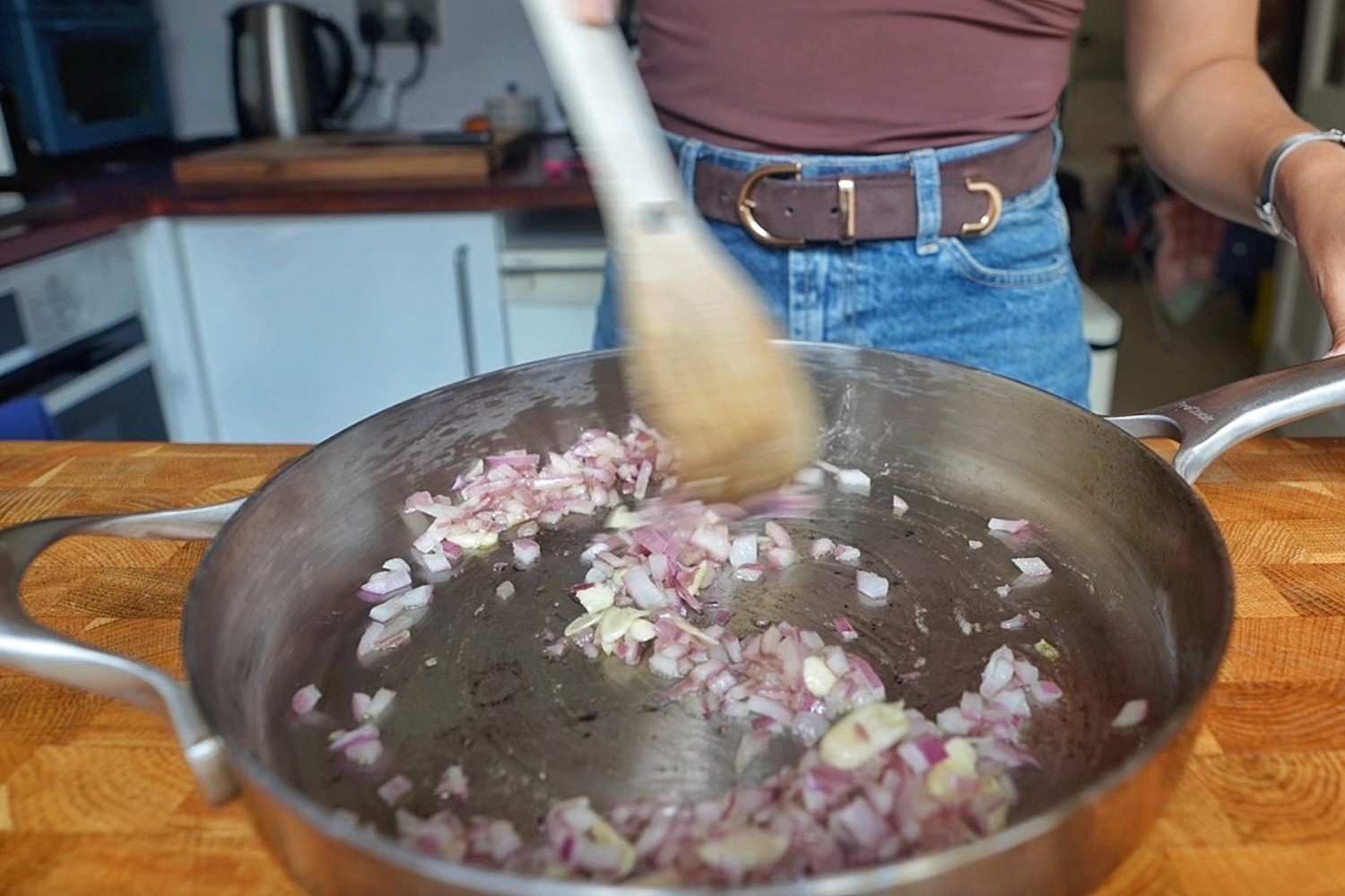 Finely diced red onion and sliced garlic frying in a stainless steel pan with a wooden spoon.
