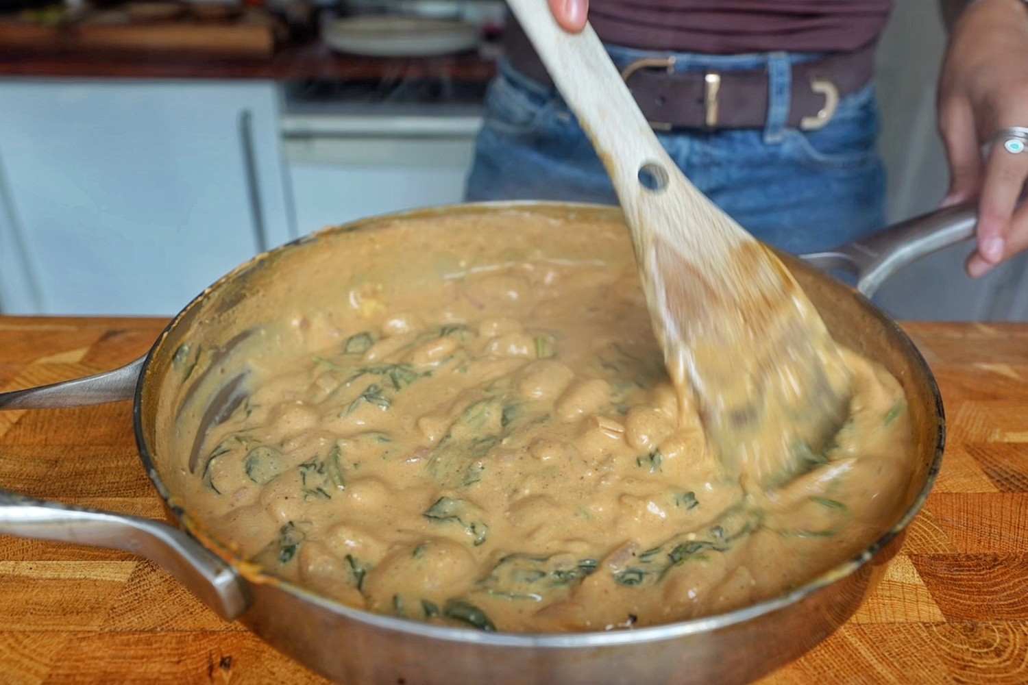 Creamy curried butter beans with wilted spinach being stirred in a stainless steel pan.