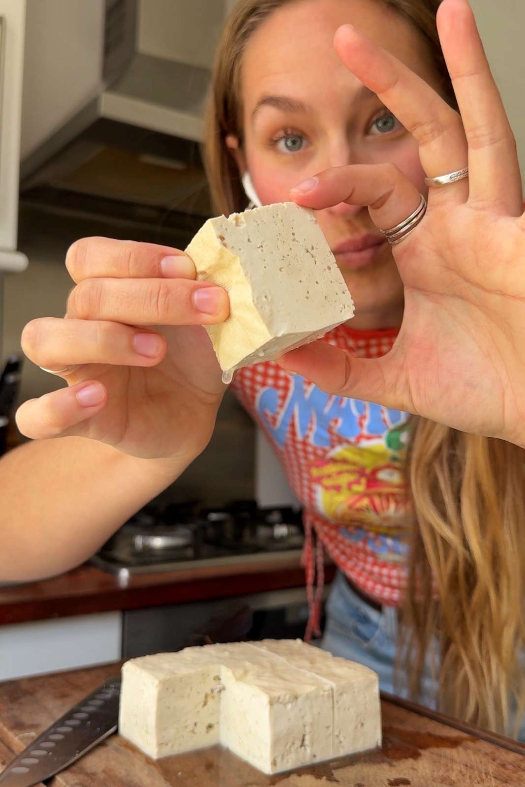 Emily Roz holding up a 2-inch cube of firm tofu, with the rest of the block cut into giant cubes on a wooden board.