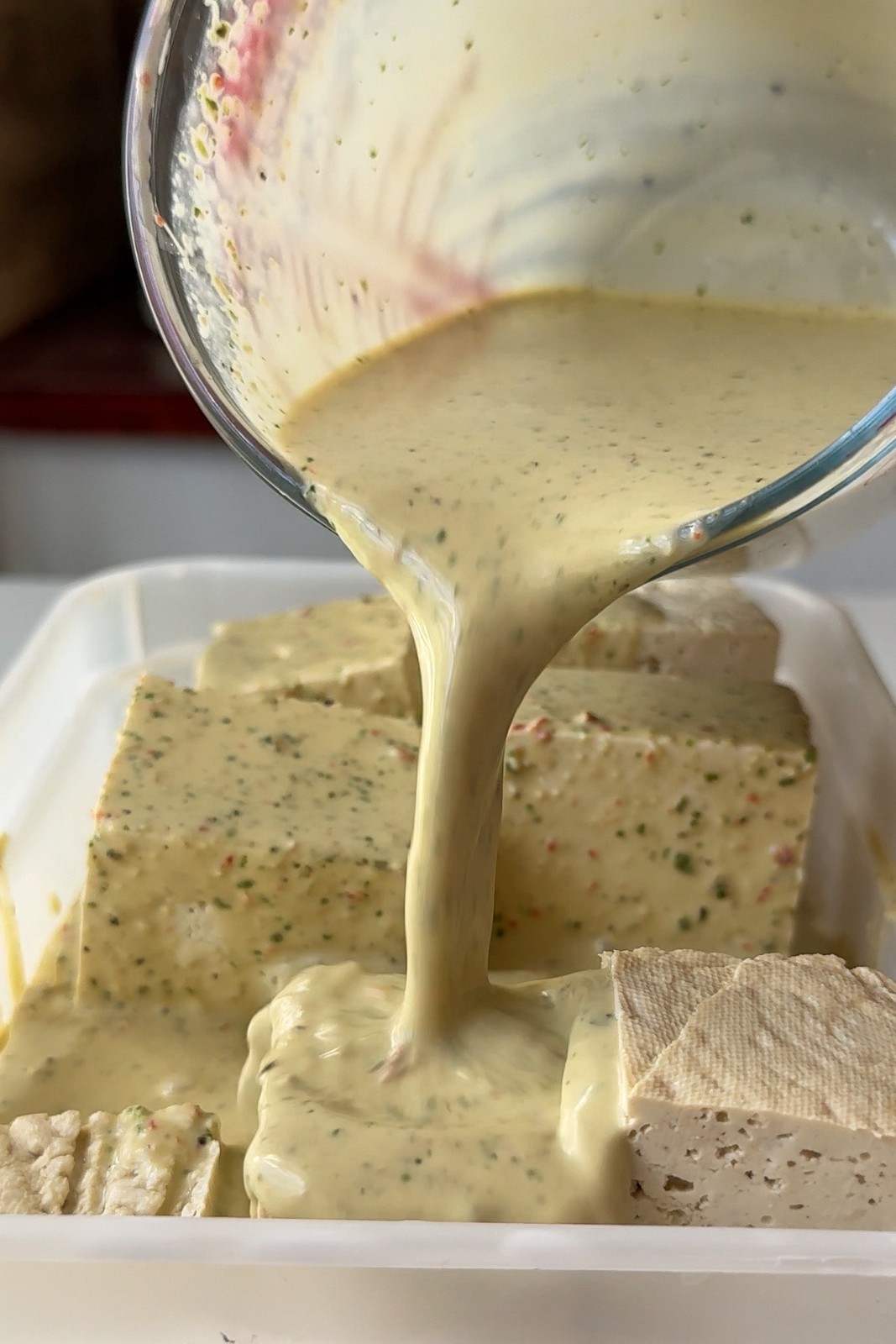 Pouring the coconut and lime leaf marinade over large pieces of firm tofu in a plastic container.