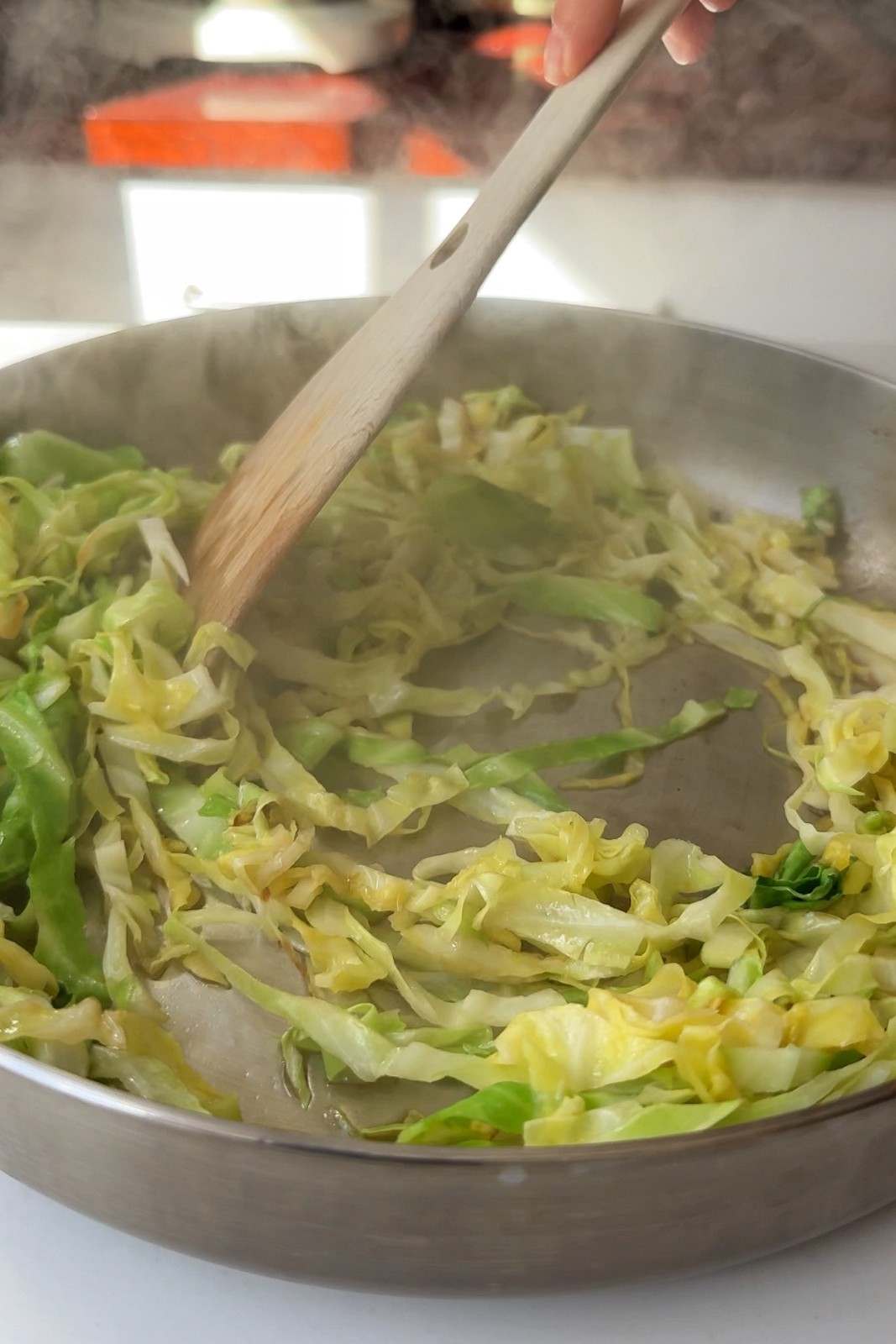 Sliced cabbage frying in a stainless steel skillet, stirred with a wooden spoon until softened.