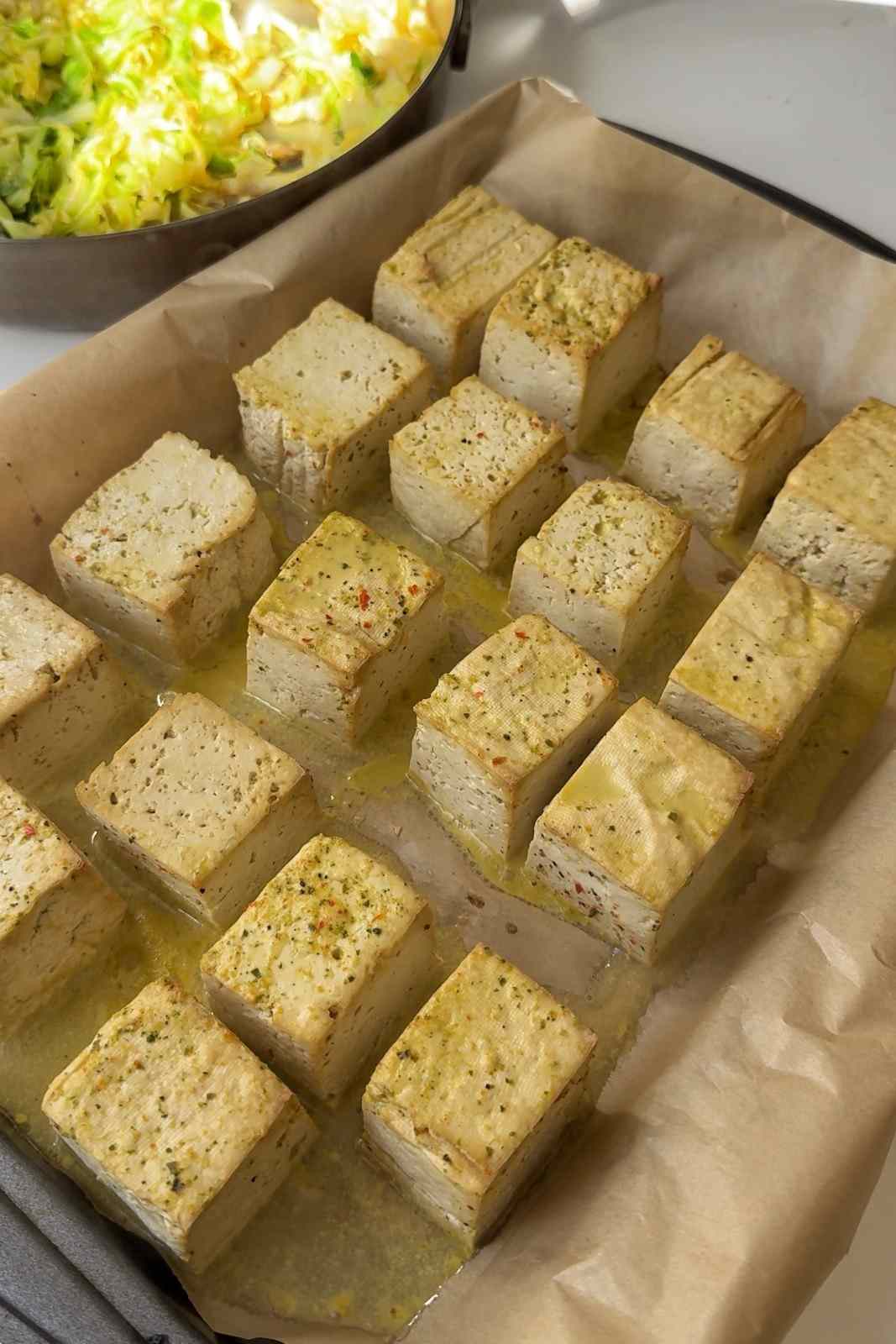 Giant tofu cubes baked until lightly golden on a parchment-lined sheet pan, with a skillet of fried cabbage behind.