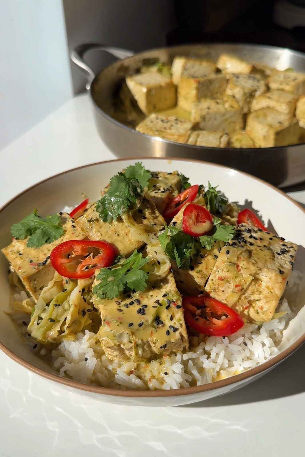 Bowl of giant tofu curry over jasmine rice in front of the skillet, garnished with cilantro, red chili and sesame seeds.