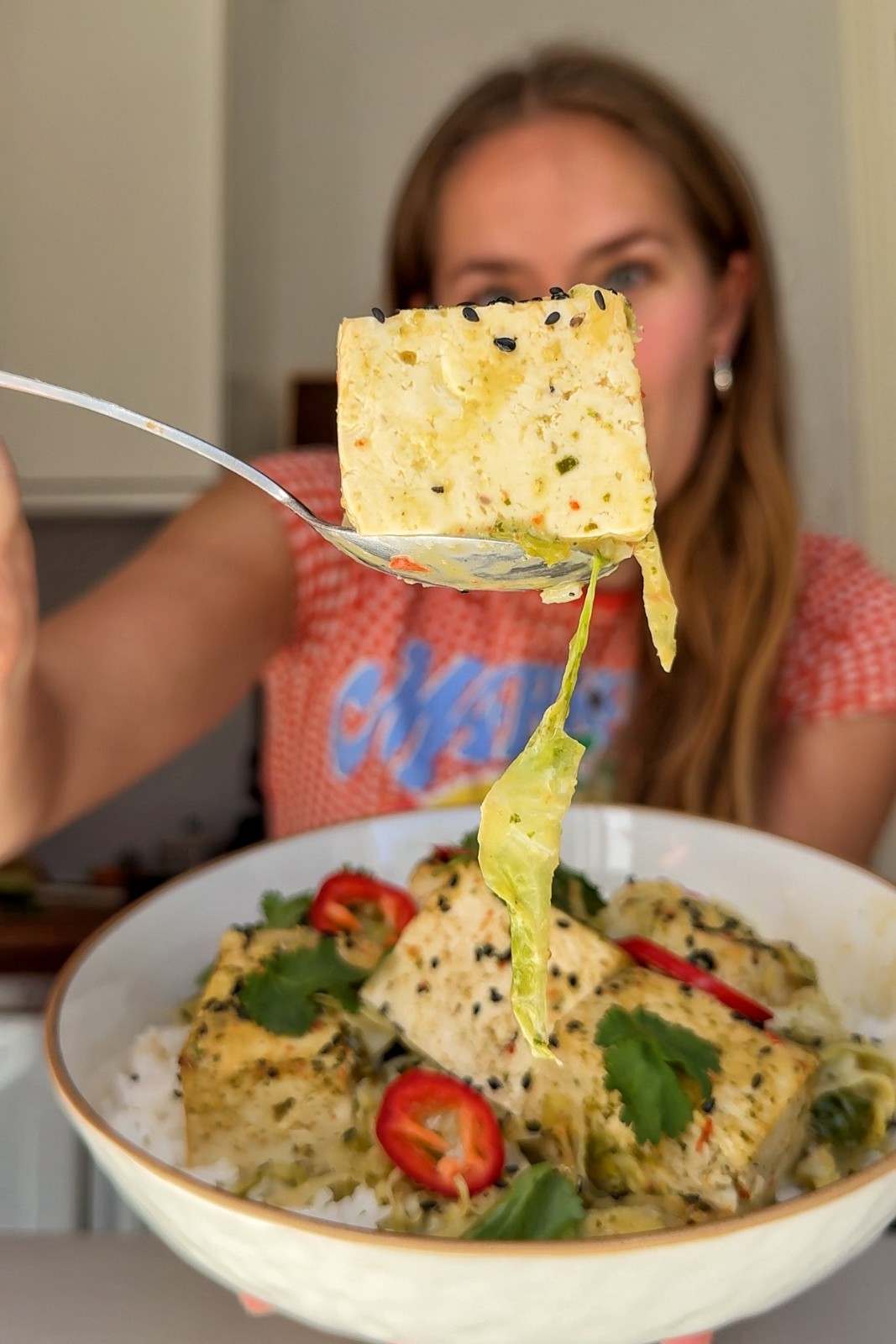 Emily Roz holding a spoonful of giant tofu curry showing a soft tofu cube and a strand of cabbage, held over a full bowl.