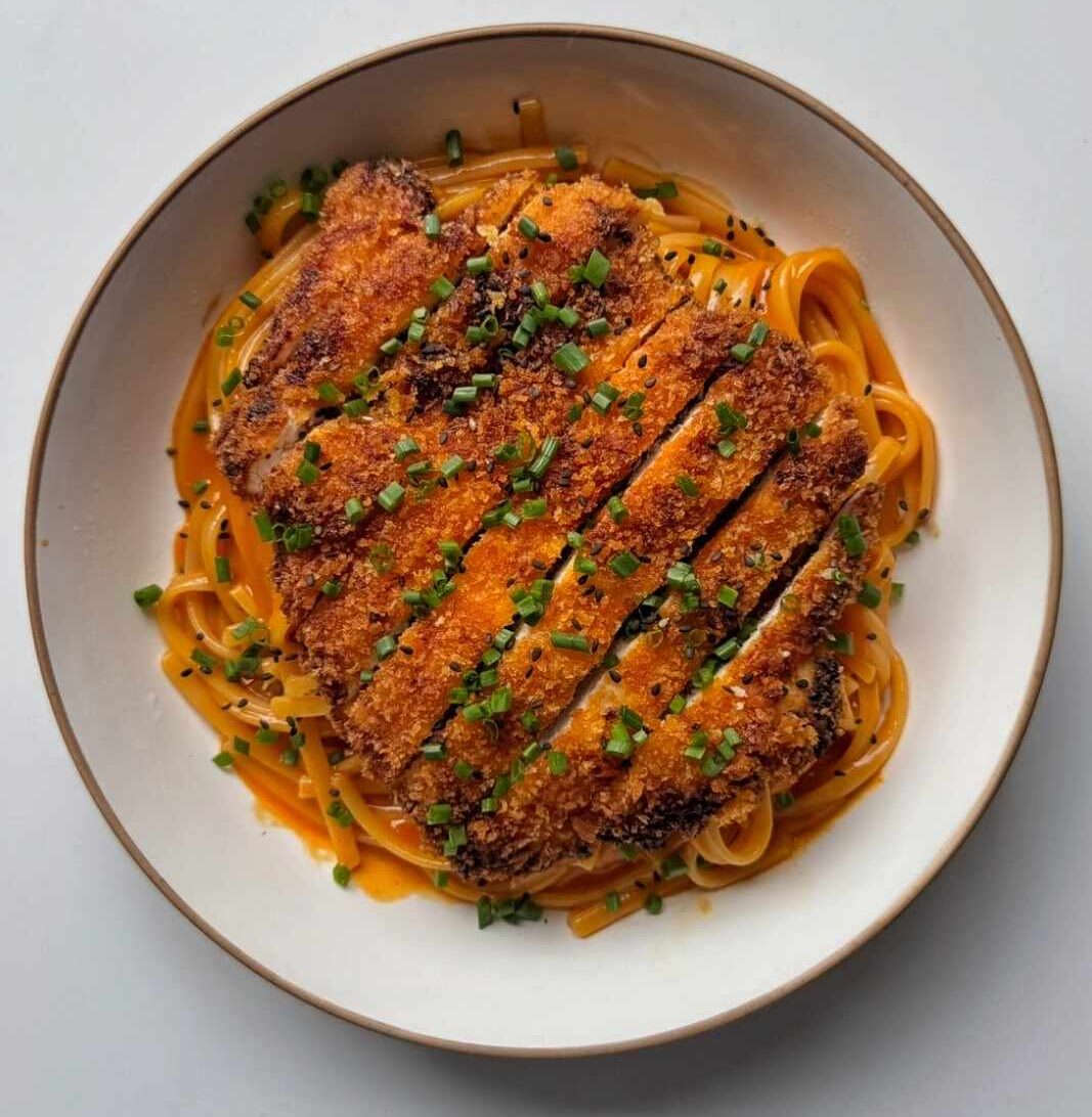 Overhead view of Korean fried chicken pasta, with sliced crispy chicken on creamy gochujang linguine in a white bowl, topped with chives and black sesame seeds.