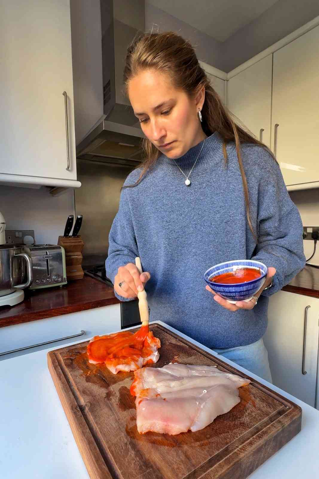 Emily Roz brushing gochujang mayo onto flattened raw chicken breasts on a wooden cutting board, holding a small blue and white bowl of the red sauce.