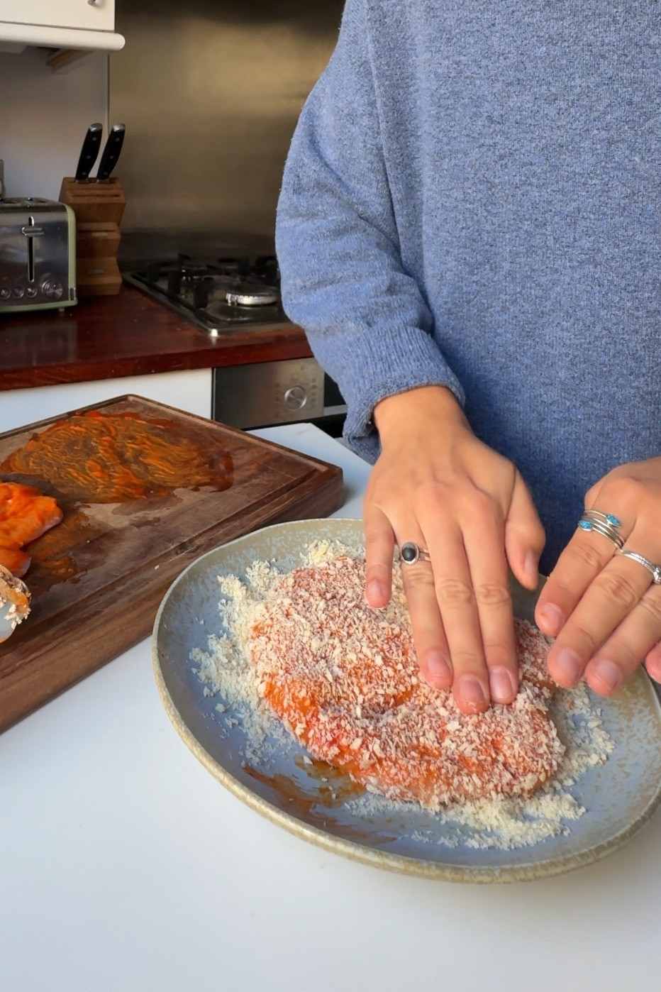 Emily Roz pressing a gochujang mayo coated chicken cutlet into panko breadcrumbs on a blue ceramic plate.