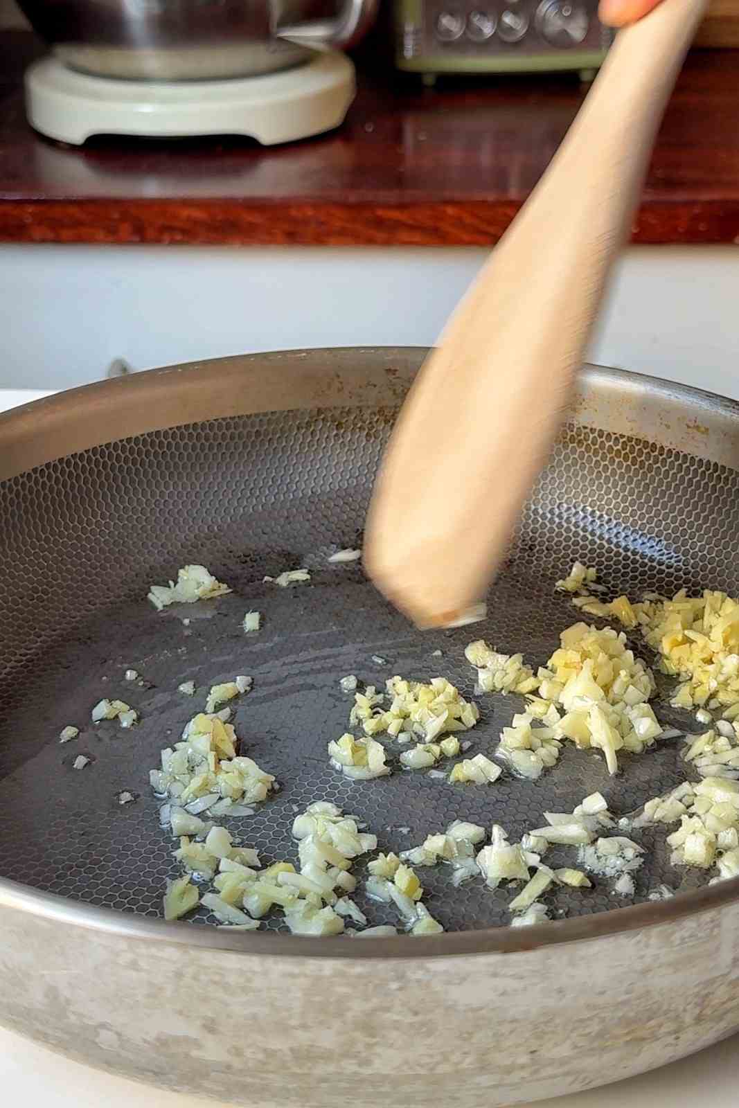 Finely chopped garlic and ginger sizzling in olive oil in a stainless steel skillet, stirred with a wooden spoon.