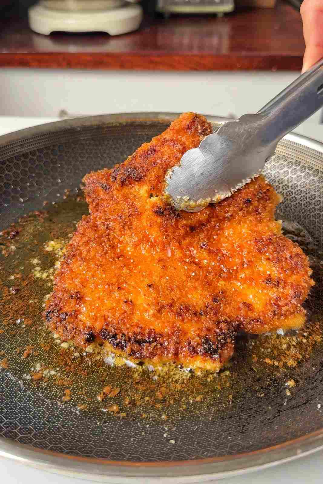 Tongs lifting a crispy panko chicken cutlet out of shallow oil in a skillet, showing its deep golden, caramelized crust.