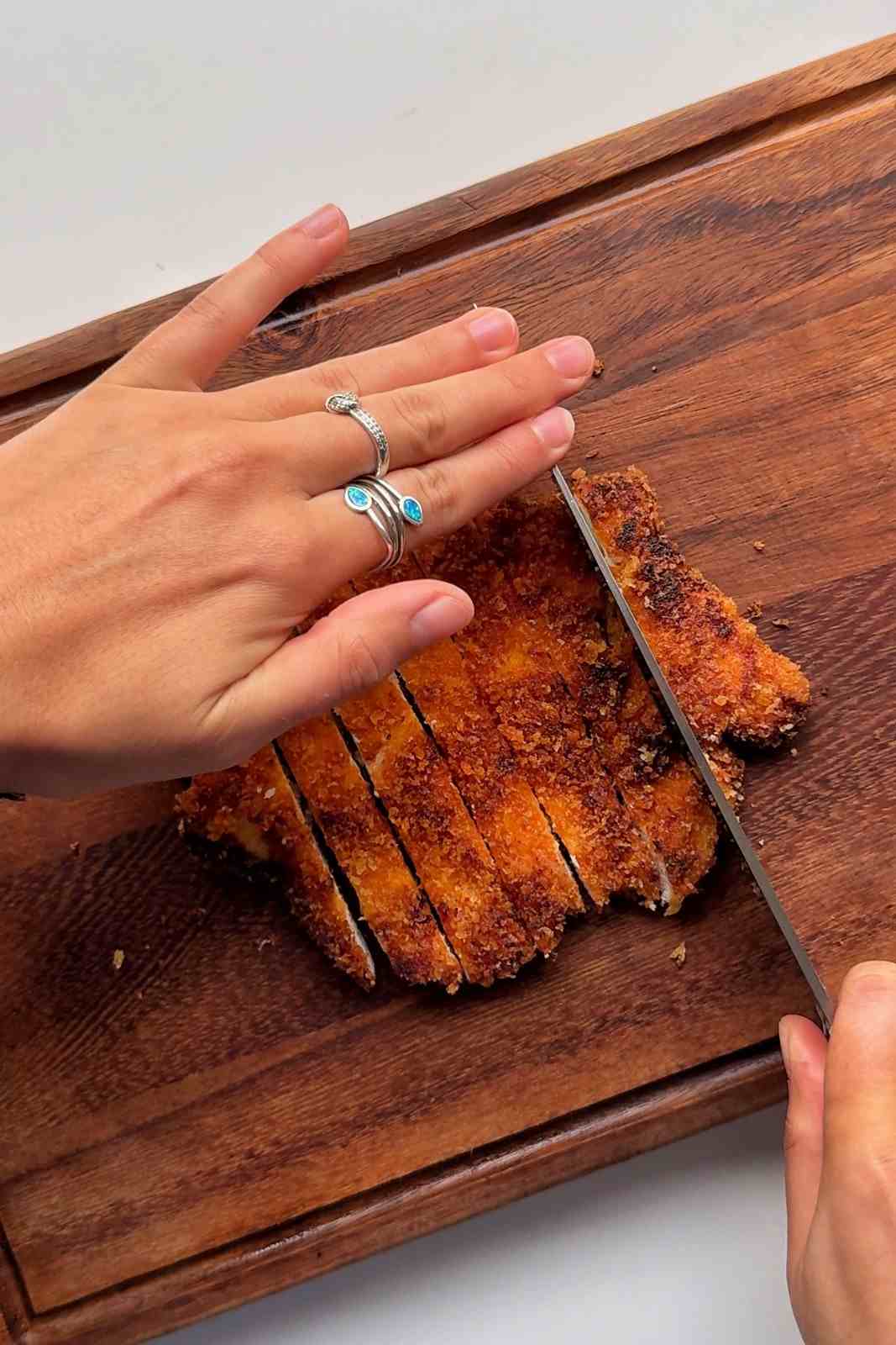 Slicing a crispy gochujang panko chicken cutlet into strips on a wooden cutting board.