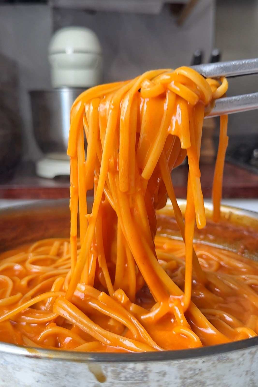Tongs lifting linguine coated in glossy, creamy gochujang sauce out of a stainless steel skillet.