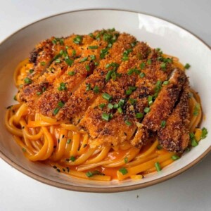 Korean fried chicken pasta in a white bowl, with a sliced golden panko chicken cutlet on creamy orange gochujang linguine, topped with chives and black sesame seeds.