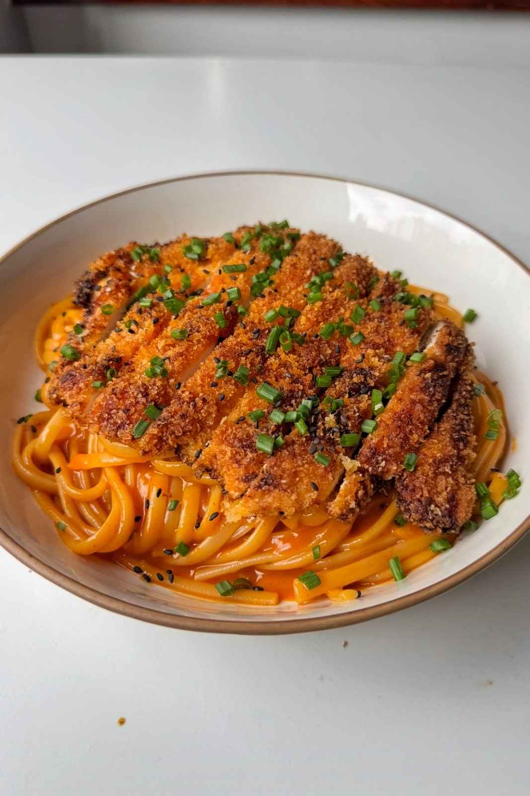 Korean fried chicken pasta in a white bowl, with a sliced golden panko chicken cutlet on creamy orange gochujang linguine, topped with chives and black sesame seeds.