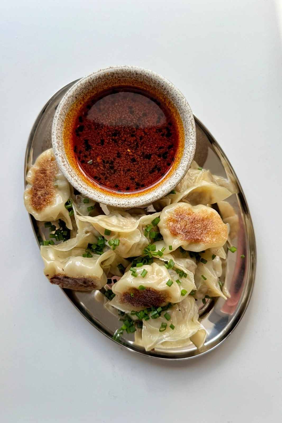 Overhead view of chicken potstickers with crispy golden bottoms on an oval metal plate, scattered with chives, next to a bowl of chili dipping sauce.