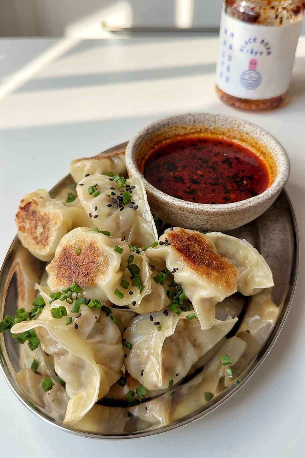 Pan-fried potstickers topped with chives and black sesame seeds on an oval plate, with a bowl of chili dipping sauce and a jar of chili oil behind.