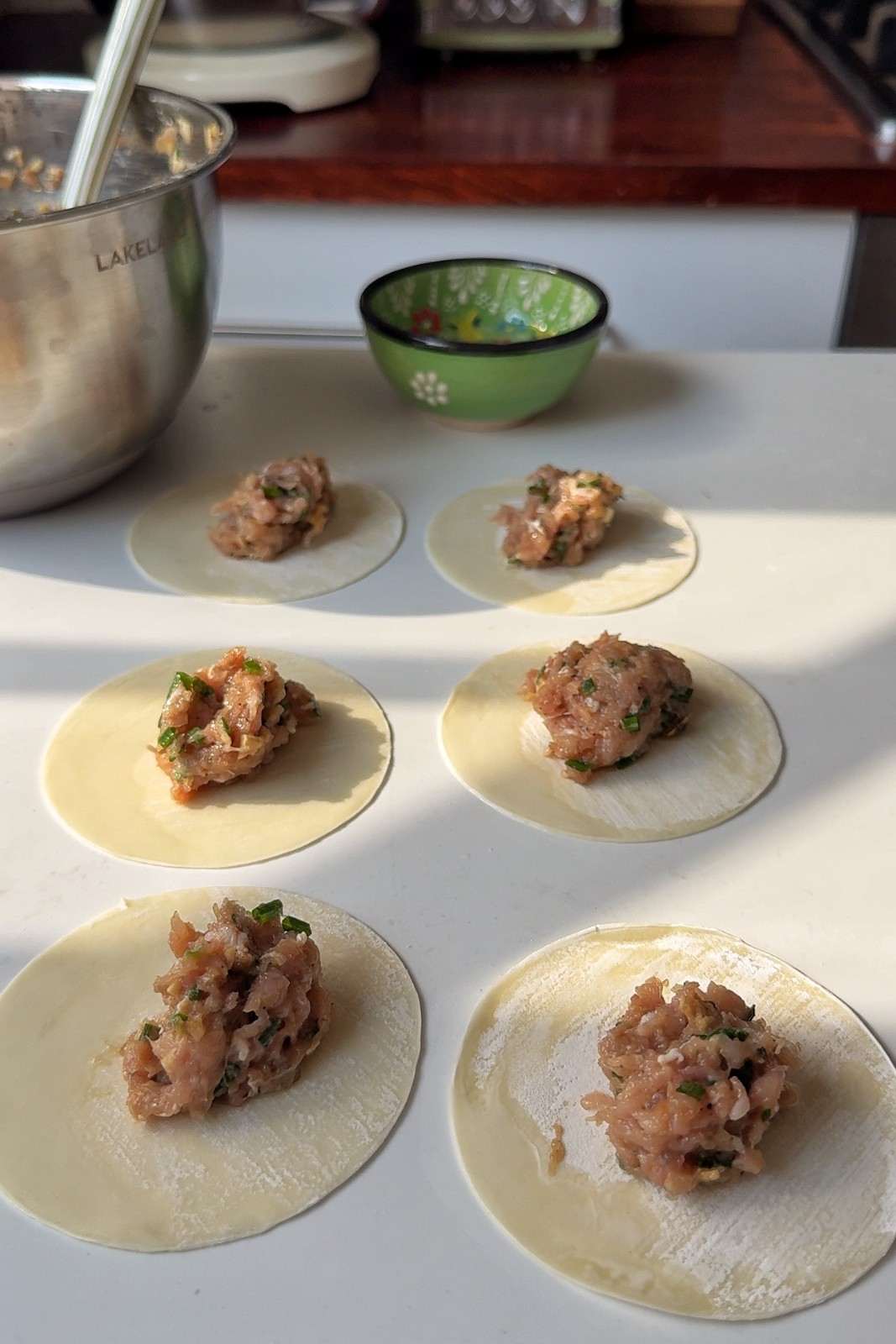 Six round dumpling wrappers on a white countertop, each with a spoonful of chicken filling in the center, beside a small bowl of water.
