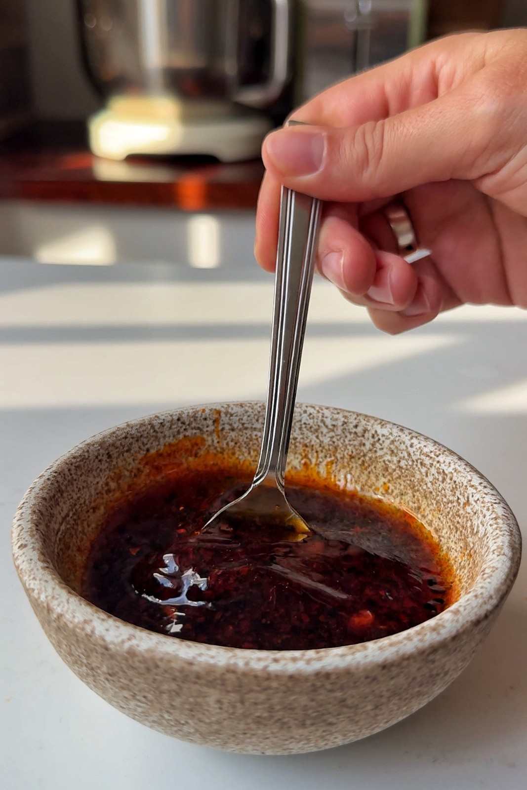Stirring a chili oil dipping sauce with a small spoon in a speckled ceramic bowl.