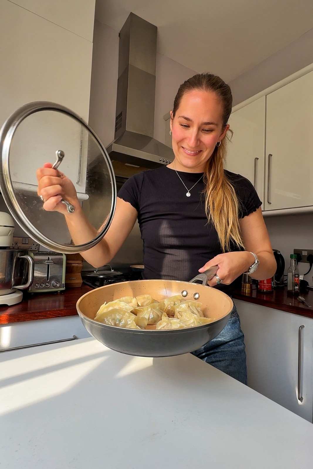 Emily Roz lifting the glass lid off a skillet of steamed potstickers in her kitchen.