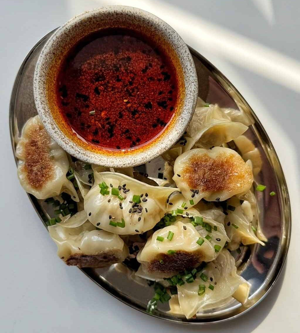 Overhead view of potstickers on an oval metal plate topped with chives and black sesame seeds, with a bowl of chili oil dipping sauce.