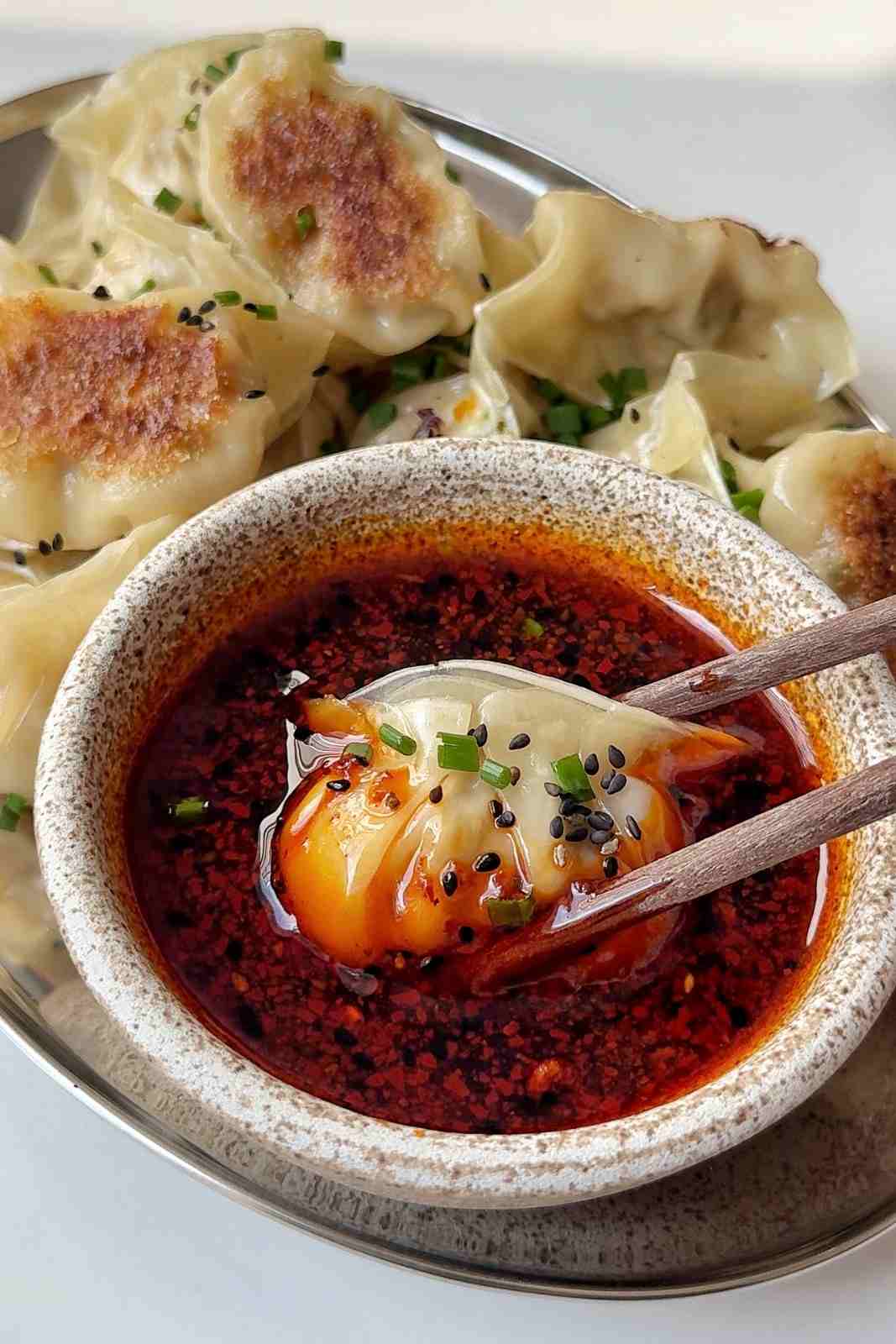 A potsticker held in chopsticks being dipped into a bowl of chili oil dipping sauce, with a plate of crispy-bottomed potstickers behind it.