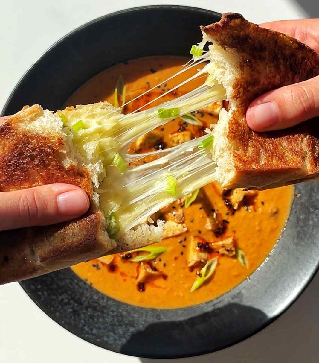 Emily Roz pulling apart a scallion cheese toastie above a bowl of tofu tomato soup.