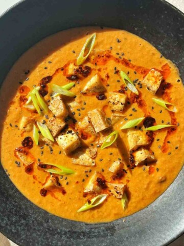 Soup toppings on a bowl of tofu tomato soup, including crispy tofu cubes, chili oil, sliced scallions, and black sesame seeds.