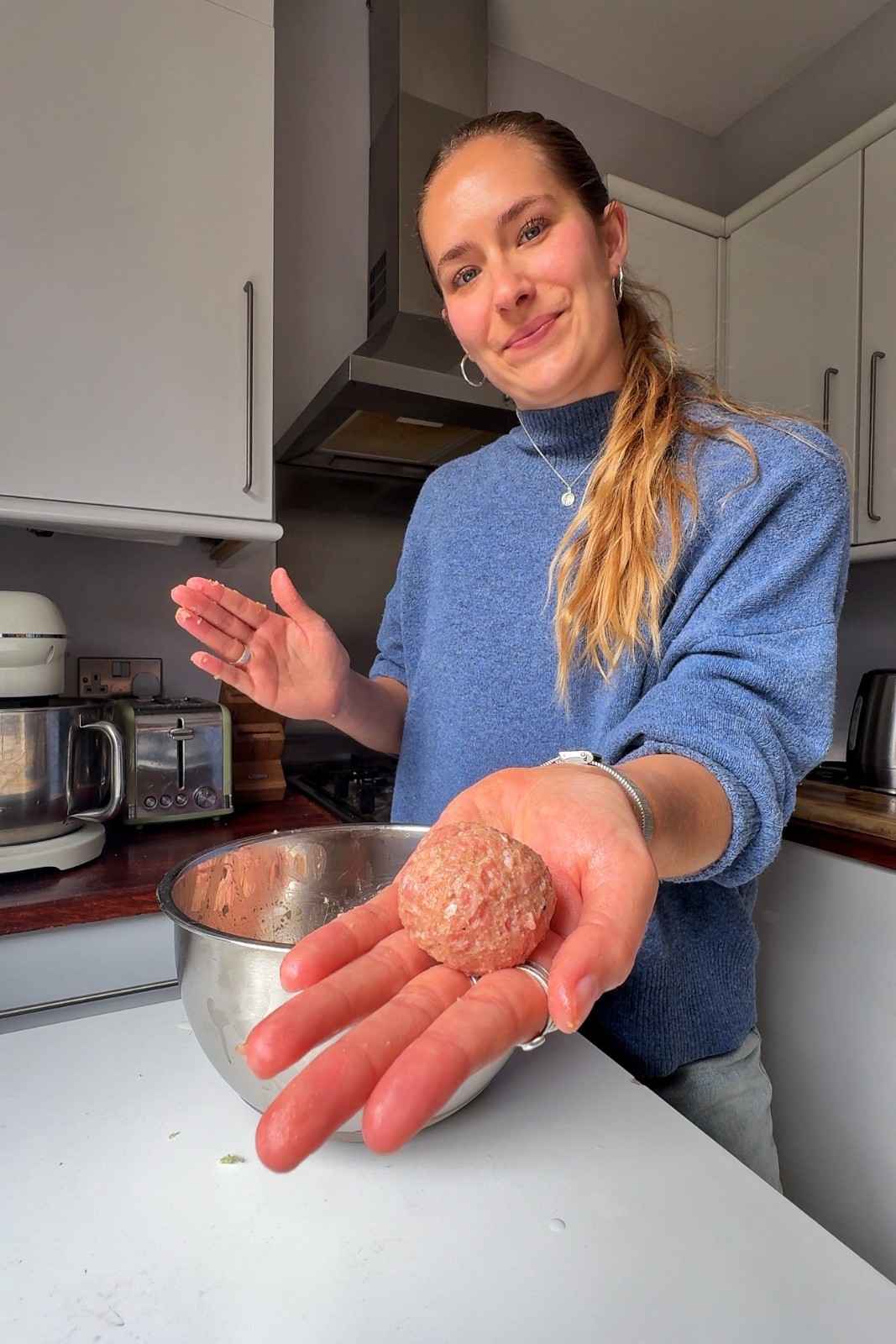 Emily holding a raw Swedish meatball in her palm, with a steel mixing bowl of the pork and beef mixture behind her.