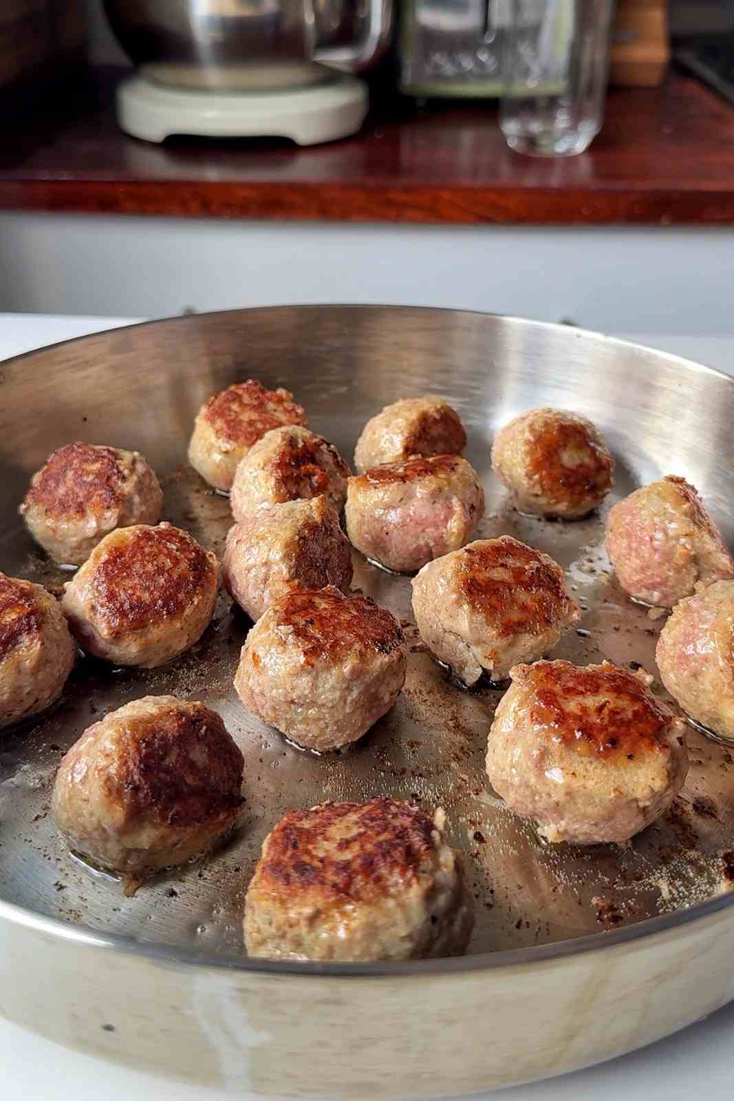 Swedish meatballs frying in a stainless steel skillet, several turned to show their deep golden crusts.