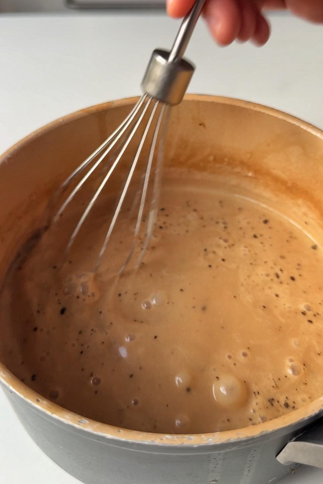 A whisk stirring creamy brown gravy flecked with black pepper as it bubbles in a saucepan.