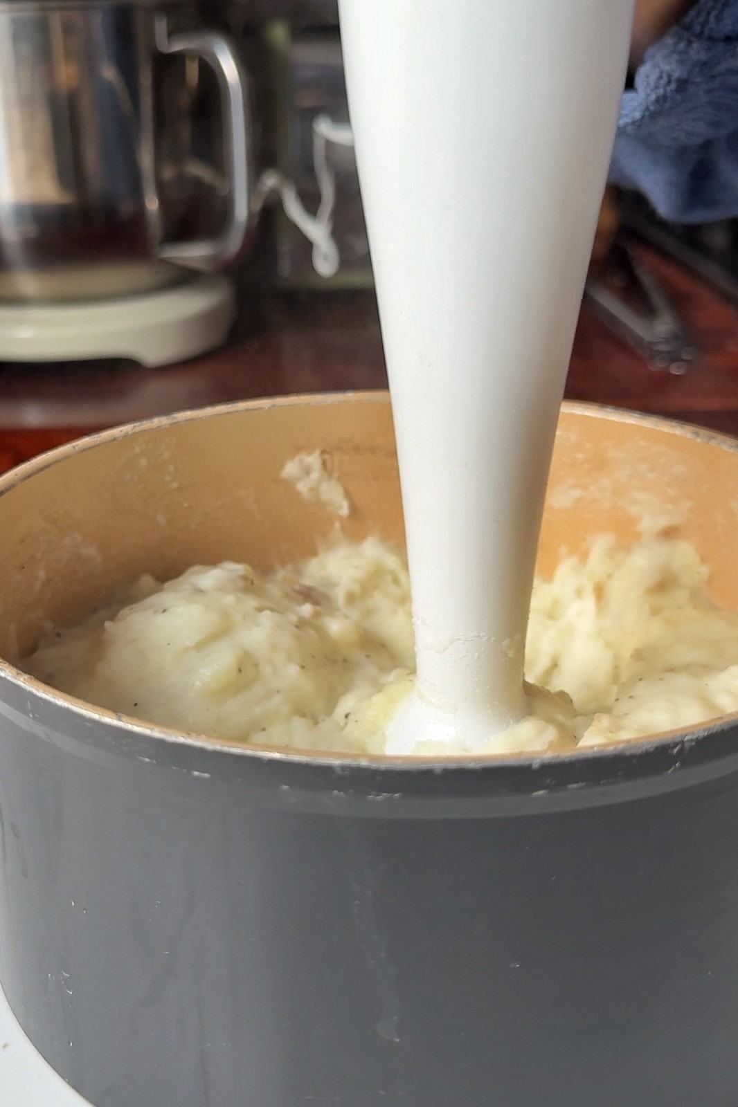 A hand blender blending boiled potatoes into smooth, pepper-flecked mashed potatoes in a saucepan.
