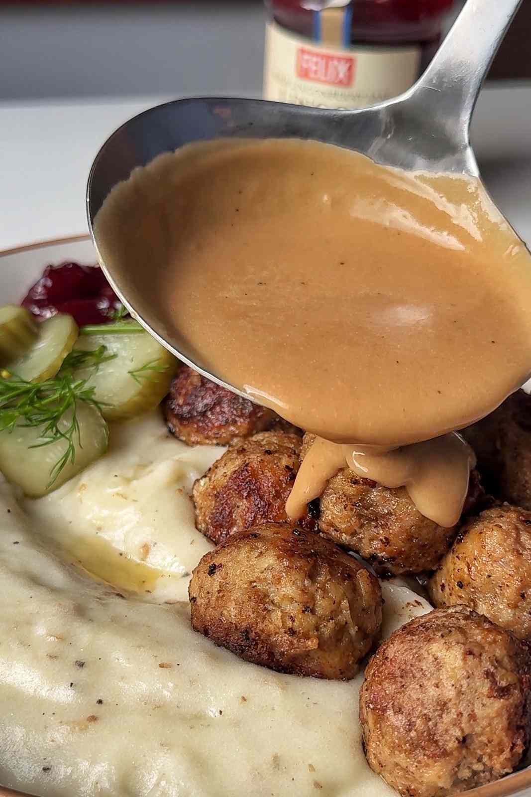 A ladle pouring creamy brown gravy over golden Swedish meatballs on mashed potatoes, with pickled cucumbers, dill and lingonberry jam.