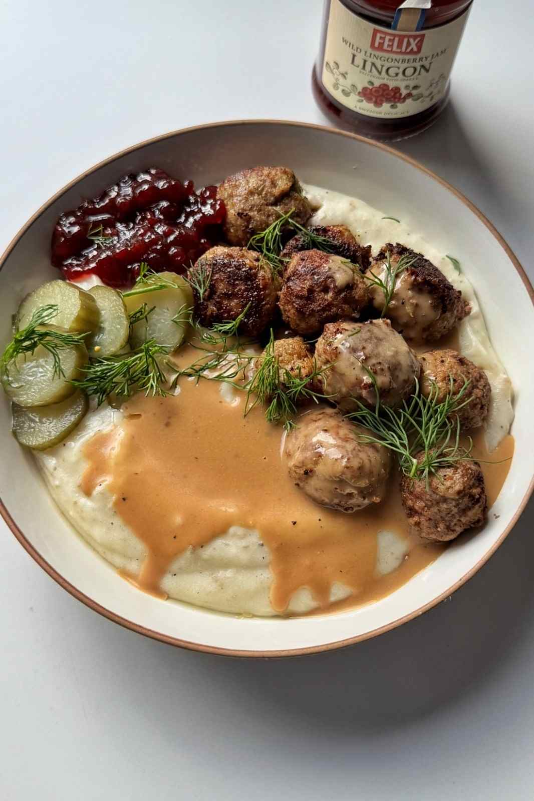 Overhead view of Swedish meatballs over mashed potatoes with creamy gravy, lingonberry jam, pickled cucumbers and dill, next to a jar of lingonberry jam.