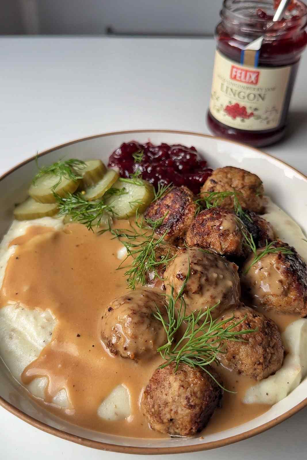 A bowl of Swedish meatballs on mashed potatoes with creamy brown gravy, lingonberry jam, pickled cucumber slices and fresh dill, with an open jar of lingonberry jam behind.