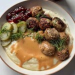 A bowl of Swedish meatballs on mashed potatoes with creamy brown gravy, lingonberry jam, pickled cucumber slices and fresh dill, with an open jar of lingonberry jam behind.